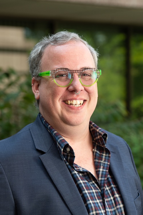 Smiling white person with short graying hair, green geometric glasses, wearing a plaid button up shirt and blue sports coat