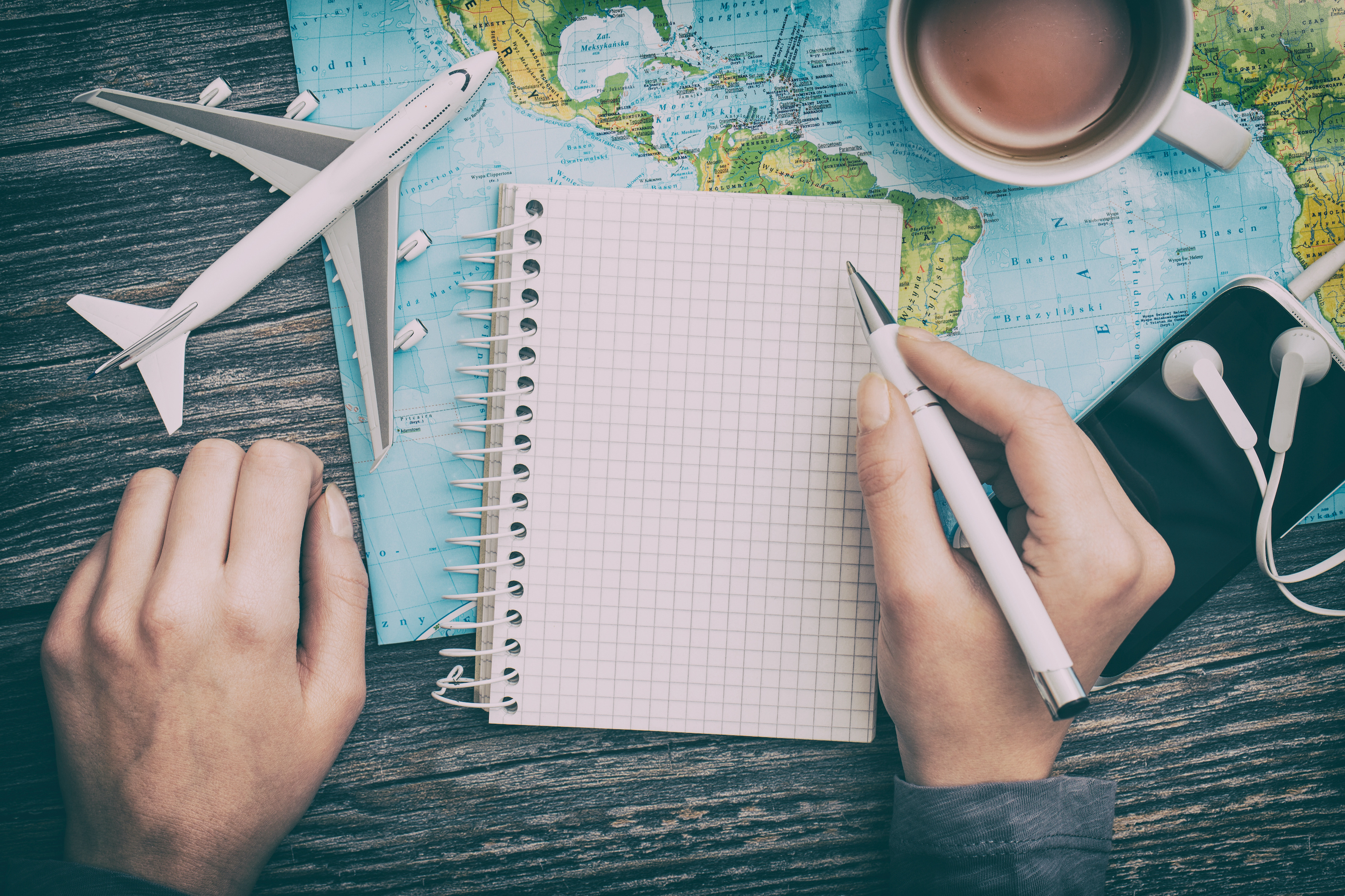 Student writing in travel journal with map, toy plane, coffee and phone