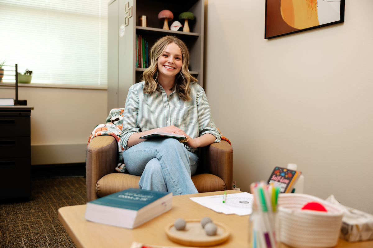 A female student therapist sitting in a chair smiling and holding a notepad.